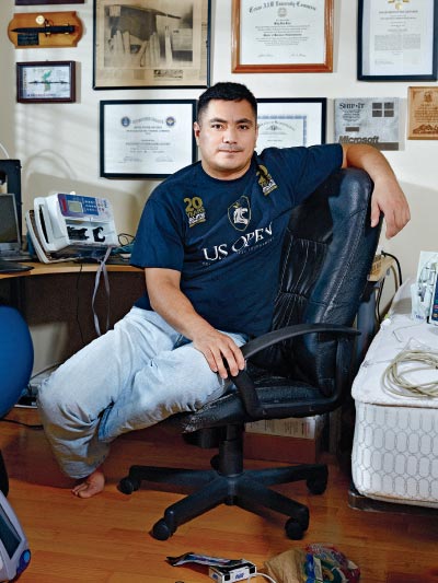 Photograph of Billy Rios, subject of this story, in a U.S. Open t-shirt and blue jeans, casual post reclining on swivel chair on hardwood floor. An exposed sheetless mattress with coiled cord atop is visible on the right side. Various framed certificates and degrees hang in the background.