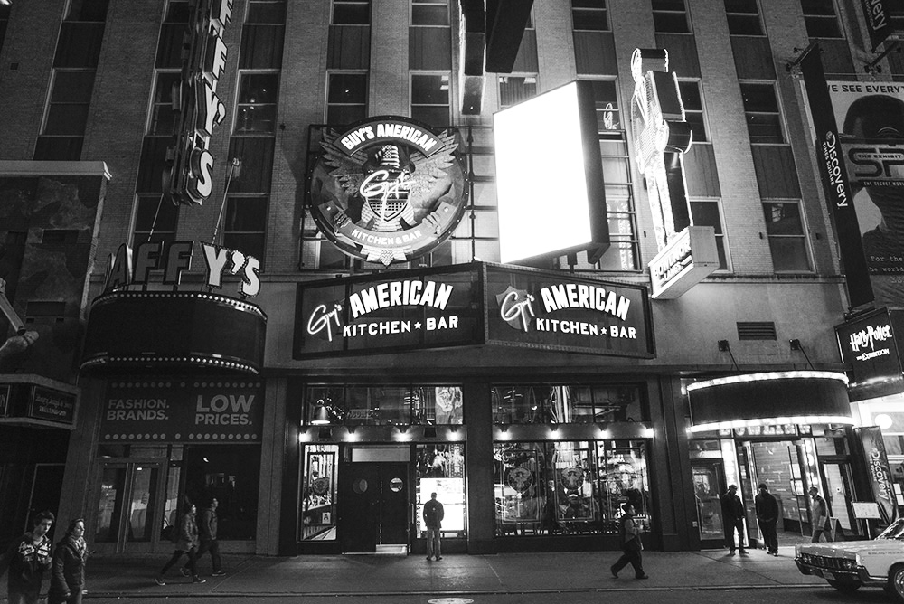 Guy Fieri’s restaurant on the exterior of the building at night, beckoning a lone tourist to the window.