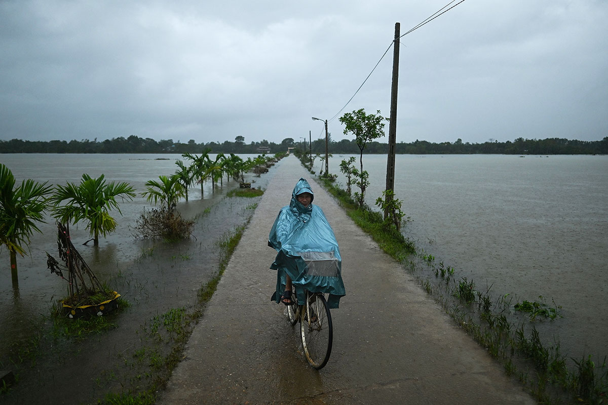 A man in a blue raincoat rides his bicycle down a raised, concrete sidewalk in the pouring rain. Fields are flooded on either side of him, with only a few stalks of vegetation poking out.